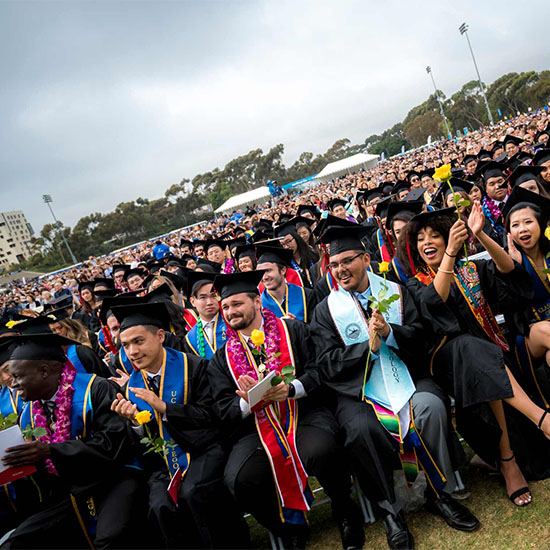 UC San Diego students at commencement - wearing caps and gowns and celebrating while sitting in the first row of a crowd of attendees